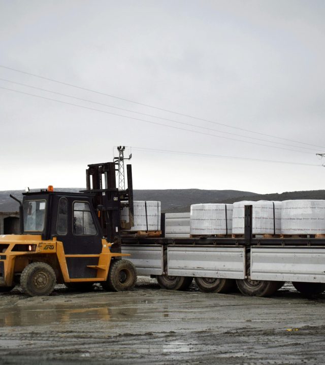A forklift is loading cargo onto a truck in a muddy logistics area with a worker assisting.